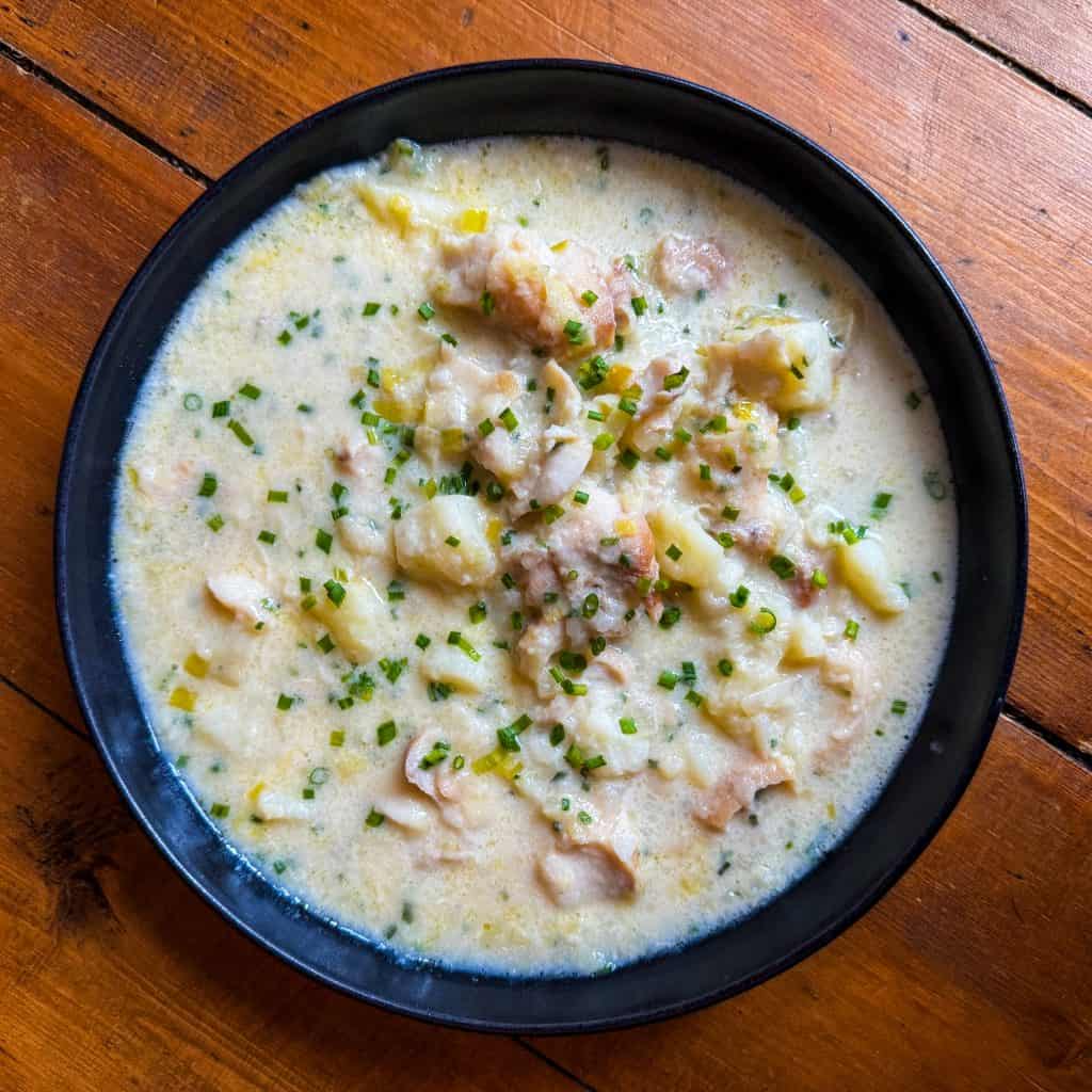 A black bowl filled with creamy clam chowder, featuring pieces of clam, potatoes, and garnished with chopped chives. The bowl is placed on a wooden table.