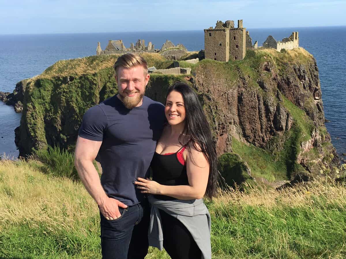 A smiling couple stands together outdoors with historic ruins of Dunnottar Castle on a grassy cliff overlooking the sea in the background on a sunny day.