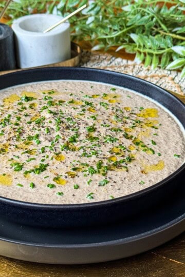 A bowl of creamy mushroom soup garnished with chopped herbs and a drizzle of olive oil sits on a decorative plate. In the background, there are small containers with utensils and a leafy green plant.