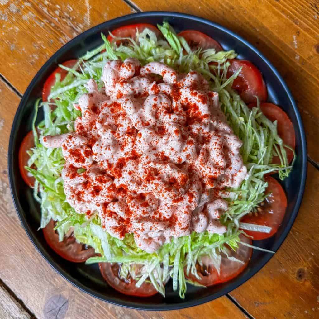 A black plate with shredded lettuce topped with a generous serving of shrimp salad, sprinkled with paprika. The dish is surrounded by slices of fresh tomato, and the plate sits on a wooden surface.