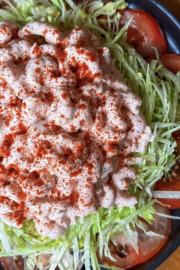 A salad with a base of shredded lettuce and sliced tomatoes. Topped with a creamy pink dressing, generously sprinkled with paprika. Served on a dark plate.