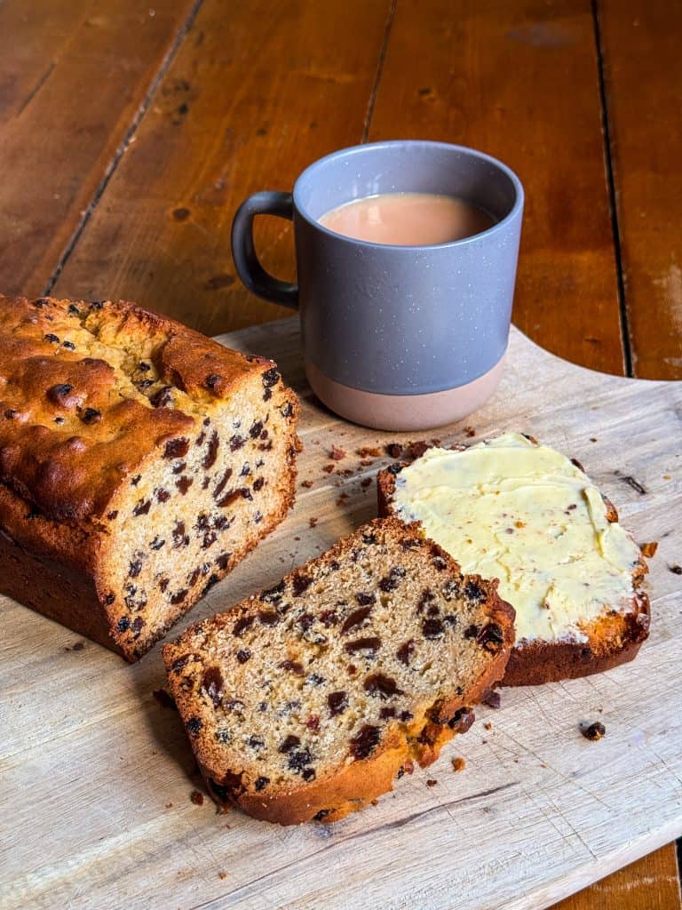 A loaf of fruit bread with two slices cut, one spread with butter, on a wooden cutting board. A dark mug filled with tea rests beside the bread, all placed on a wooden table.