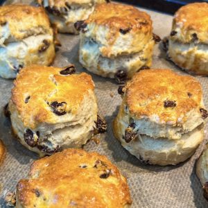 Golden-brown scones with raisins, fresh out of the oven, are placed on a parchment-lined baking tray. They have a flaky, buttery texture with a shiny, slightly crispy top.