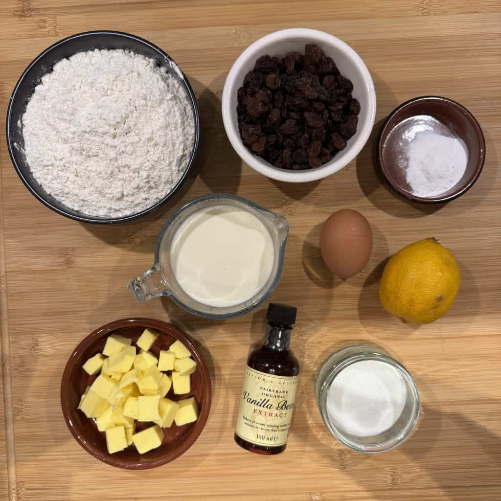 A wooden surface with ingredients for baking: a bowl of flour, raisins in a bowl, a small bowl of baking powder, a whole egg, a lemon, cream in a measuring cup, cubed butter, sugar in a cup, and a bottle of vanilla extract.