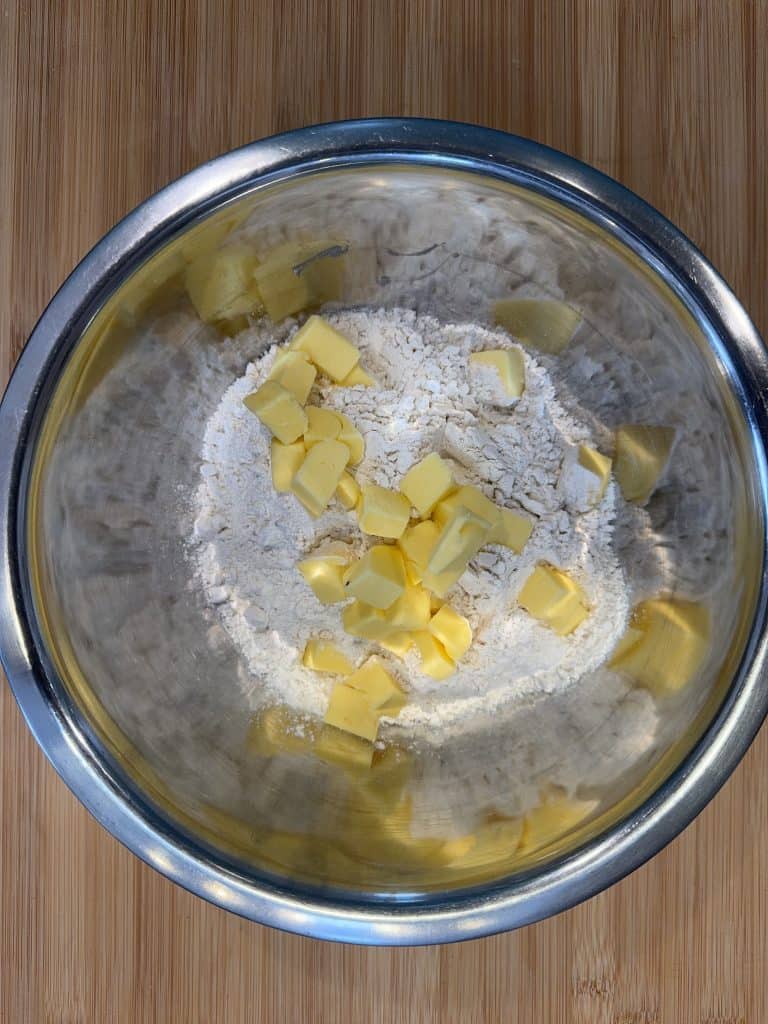 A metal mixing bowl on a wooden surface contains flour and cubes of butter. The ingredients are ready to be mixed for baking.