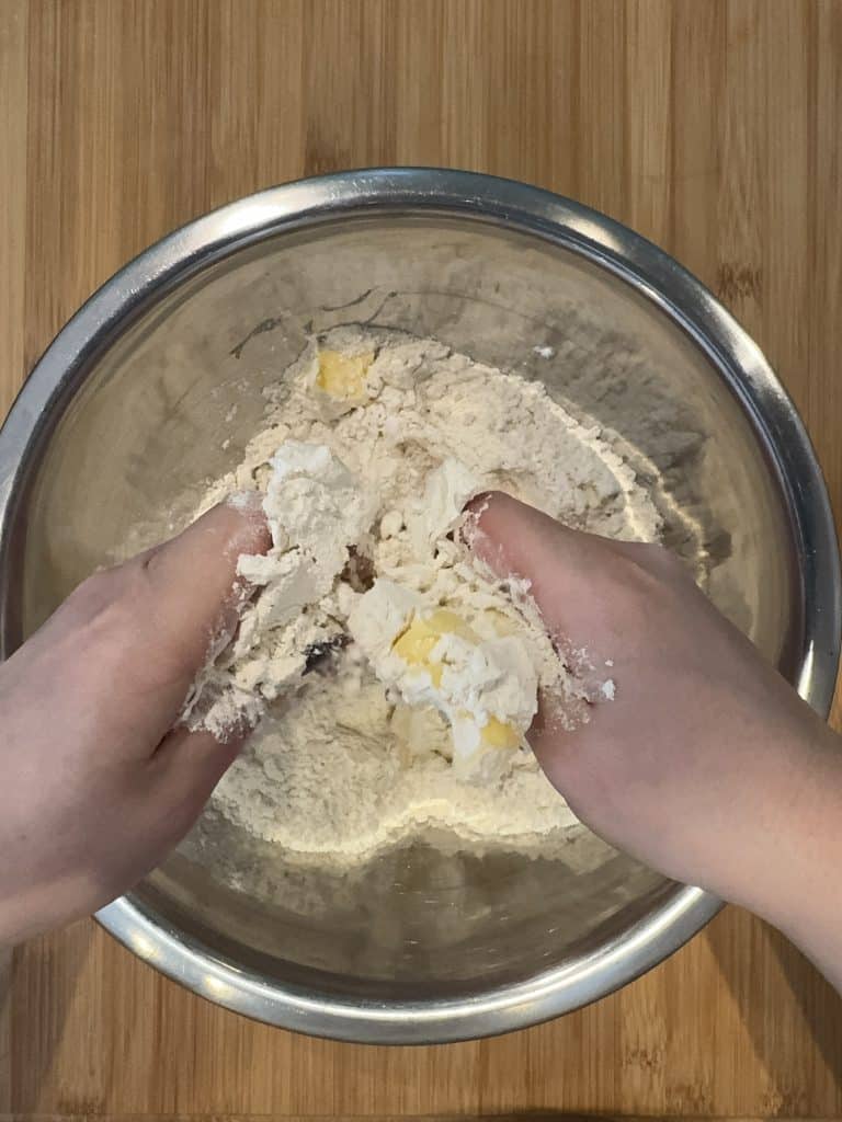 Hands mixing flour and butter in a silver bowl on a wooden surface, preparing dough.