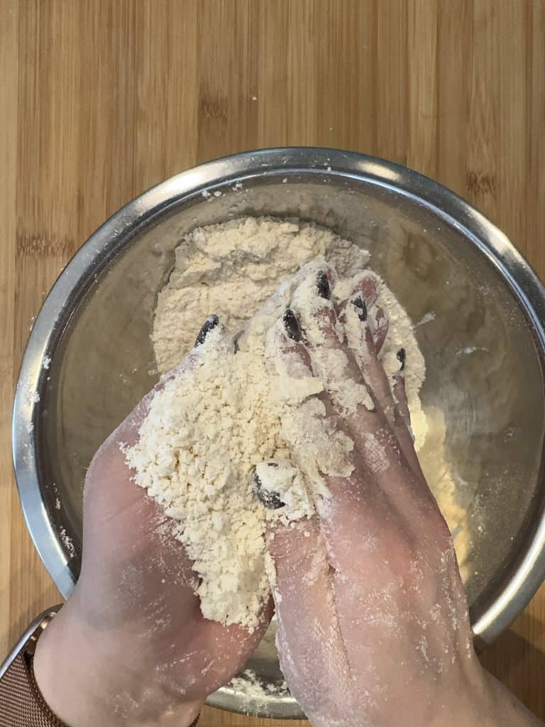 Hands are mixing flour in a metal bowl on a wooden surface. The fingernails are painted dark.