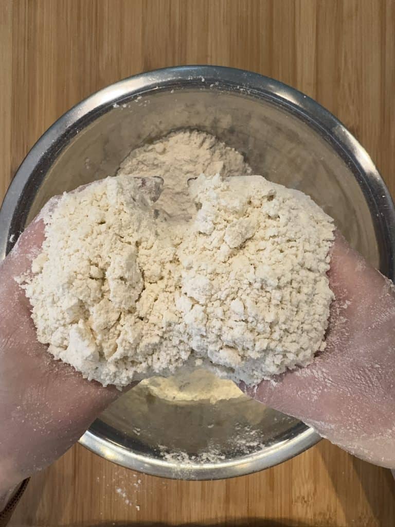 Hands mixing crumbly dough over a metal bowl on a wooden surface.