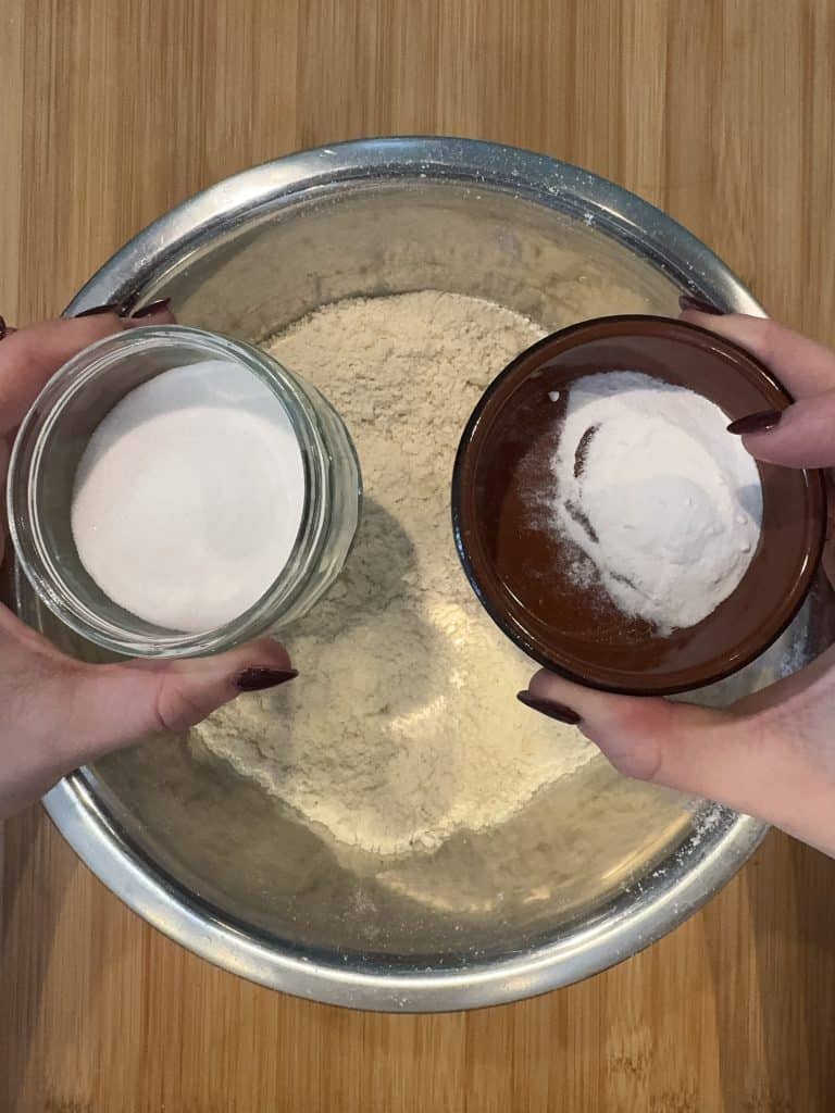Hands holding small bowls of sugar and baking powder above a larger bowl of flour on a wooden surface, preparing ingredients for baking.