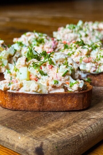 Open-faced sandwiches on toasted bread topped with a creamy mixture of diced vegetables and herbs, garnished with fresh dill, are displayed on a wooden board. The surface beneath is a warm-toned wood.