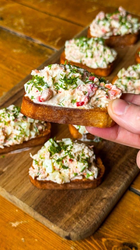 Close-up of a hand holding a piece of toasted bread topped with a creamy crab salad garnished with herbs. More pieces are on a wooden board in the background.