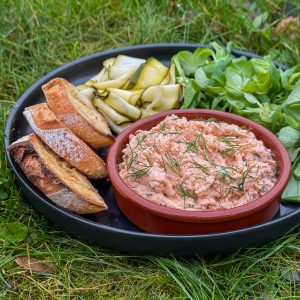 A black plate on grass holds a clay dish of salmon spread, garnished with dill. Its accompanied by toasted baguette slices, ribboned zucchini, and fresh greens.