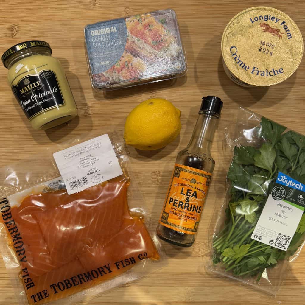 A wooden surface displays a jar of mustard, a package of soft cheese, a tub of crème fraîche, a lemon, a bottle of Lea & Perrins Worcestershire sauce, a package of parsley, and a wrapped piece of Tobermory Fish Co. salmon.