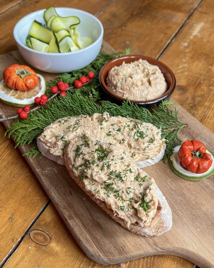 A wooden board with two slices of bread topped with a creamy spread and herbs. Nearby, a bowl of cucumber ribbons, two small tomatoes on cucumber slices, red berries, and green pine needles create a festive presentation.