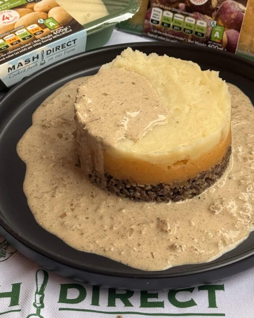 A serving of haggis, neeps, and tatties topped with creamy peppercorn sauce sits on a black plate, with extra sauce pooling around it. Packaged food and vegetables are visible in the background.