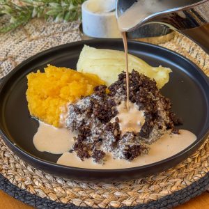 A plate with mashed sweet potatoes, mashed potatoes, and haggis topped with a creamy sauce being poured. The dish is presented on a black plate set on a woven placemat.