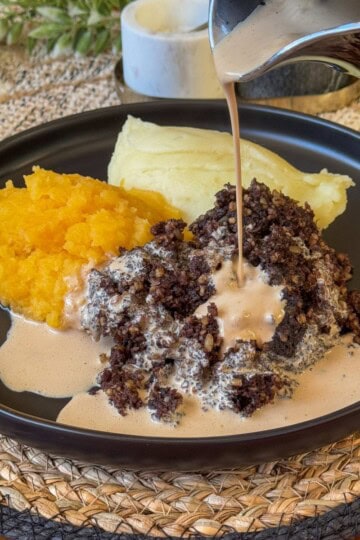 A plate with mashed sweet potatoes, mashed potatoes, and haggis topped with a creamy sauce being poured. The dish is presented on a black plate set on a woven placemat.