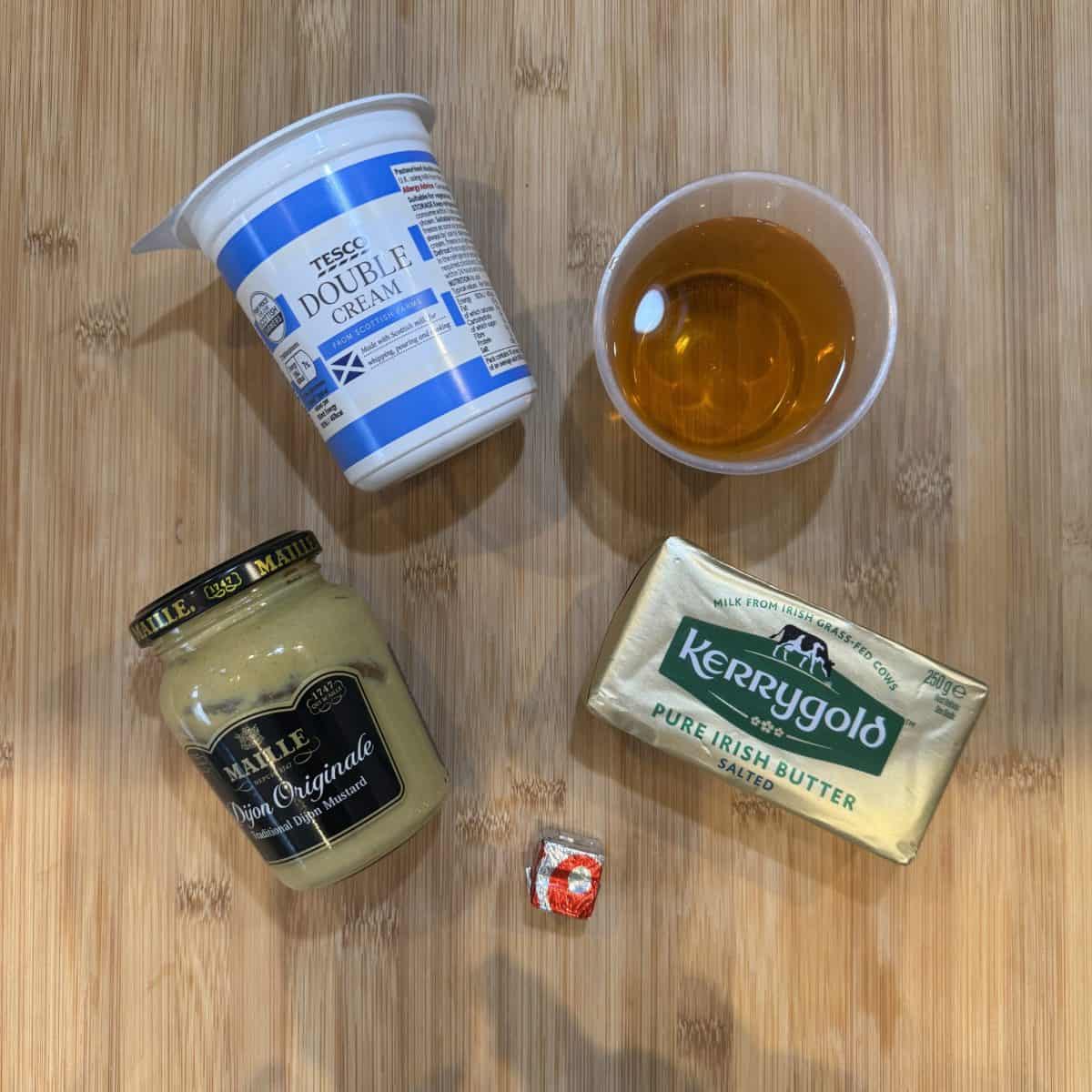 A container of double cream, a plastic cup with a golden liquid, a jar of Dijon mustard, a wrapped square of soft cheese, and a block of Irish butter are arranged on a wooden surface.