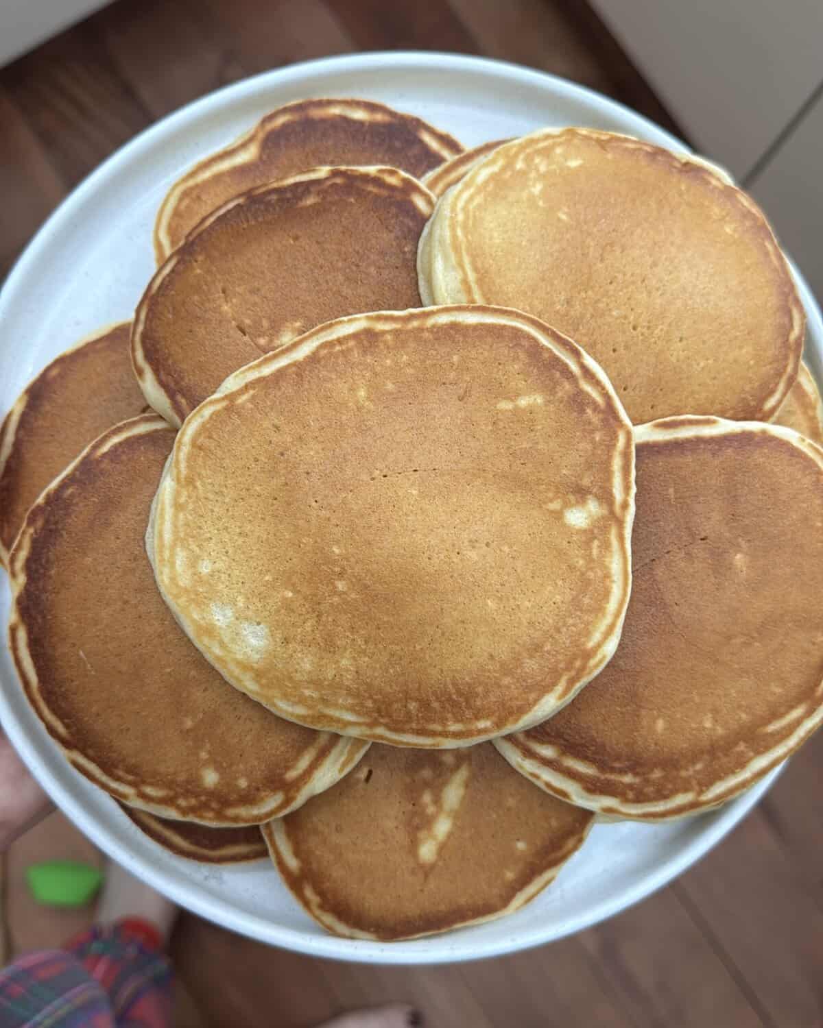 A plate piled with golden-brown self raising flour pancakes, viewed from above, resting on a wooden floor. A persons hand is holding the plate, and part of their plaid clothing and green slipper are visible at the bottom.