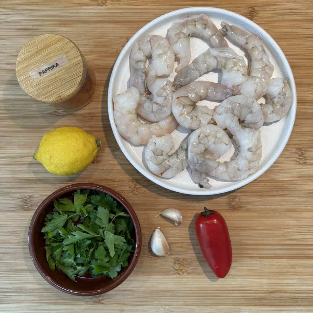 A wooden surface with a plate of raw shrimp, a jar labeled Paprika, a lemon, fresh parsley in a bowl, two garlic cloves, and a red pepper.