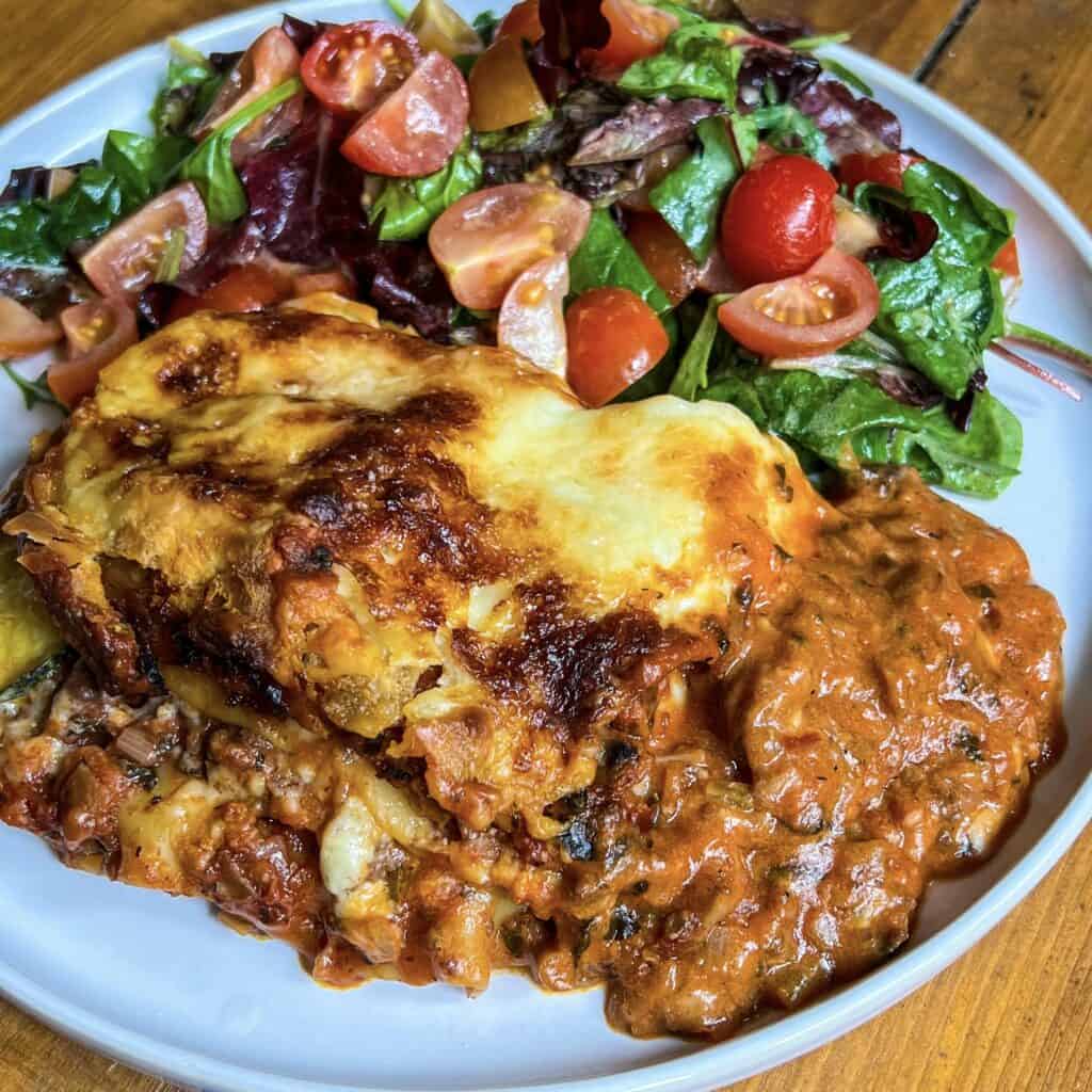 A plate of lasagne with a rich, browned cheese topping sits next to a fresh salad containing mixed greens, cherry tomatoes, and sliced radishes. The lasagna appears saucy and layered, and the salad is vibrant and colorful.
