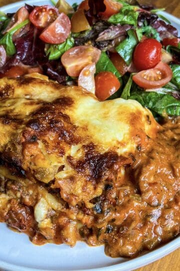 A plate of lasagna with a rich, browned cheese topping sits next to a fresh salad containing mixed greens, cherry tomatoes, and sliced radishes. The lasagna appears saucy and layered, and the salad is vibrant and colorful.
