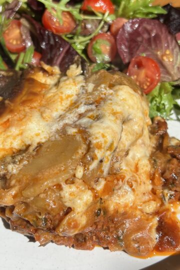 A slice of moussaka topped with béchamel sits on a white plate. Its accompanied by a fresh salad with leafy greens and cherry tomatoes. The lasagna appears rich and hearty, surrounded by a glossy sauce.