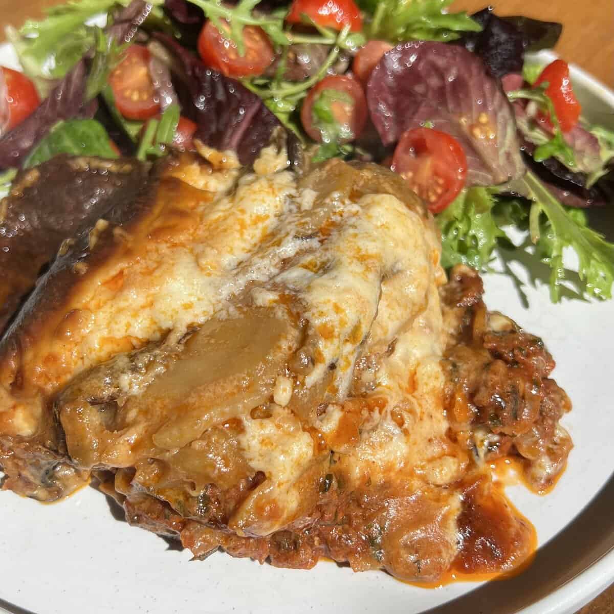 A slice of moussaka topped with béchamel sits on a white plate. Its accompanied by a fresh salad with leafy greens and cherry tomatoes. The lasagna appears rich and hearty, surrounded by a glossy sauce.