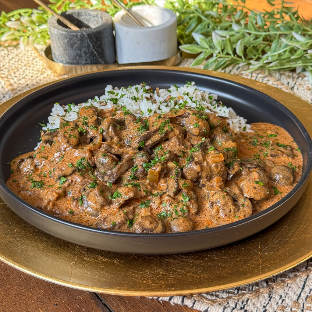 A black plate with creamy beef and mushroom stroganoff garnished with chopped herbs, served alongside rice. The dish is on a wooden table with a gold placemat, salt and pepper shakers, and lush greenery in the background.