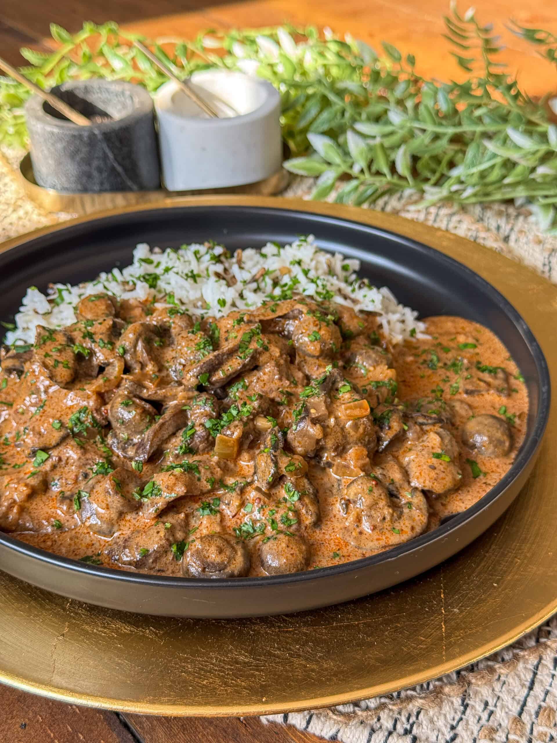 A black plate filled with creamy mushroom stroganoff garnished with chopped herbs, served alongside rice. The plate sits on a golden charger. In the background, there are plants and two containers, possibly for spices, on a wooden surface.