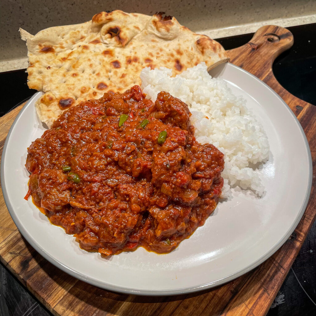 A plate with white rice, a serving of red curry garnished with herbs, and a piece of naan bread on a wooden board.