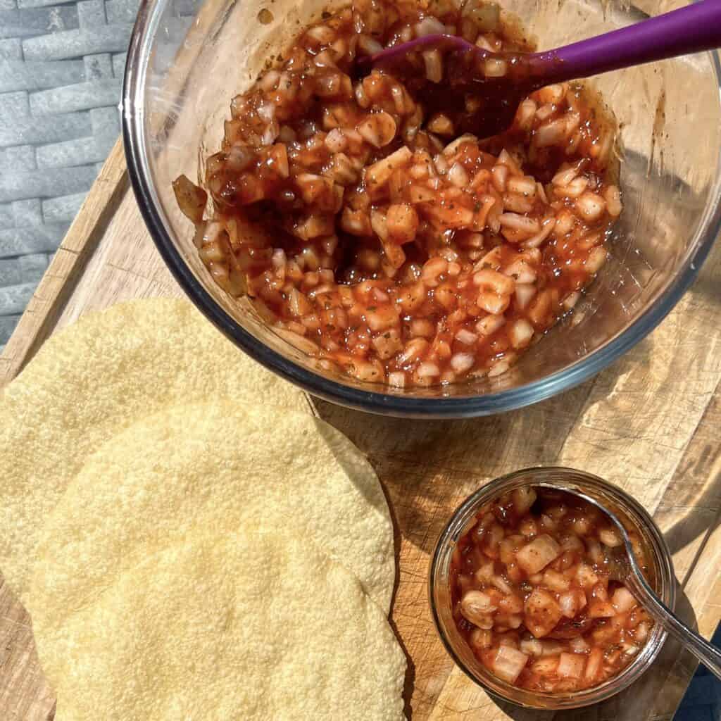 A bowl of chutney with diced onions and reddish sauce, beside two round, crispy papadums on a wooden board. A smaller jar filled with the same chutney is next to the bowl. A purple spoon is in the bowl.