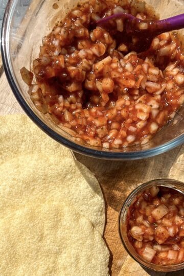 A bowl of chutney with diced onions and reddish sauce, beside two round, crispy papadums on a wooden board. A smaller jar filled with the same chutney is next to the bowl. A purple spoon is in the bowl.