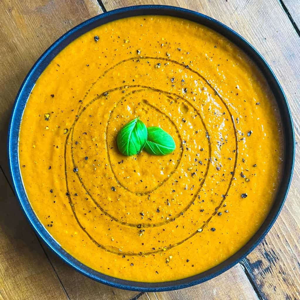 A bowl of creamy orange tomato soup garnished with a swirl of olive oil, black pepper, and two fresh basil leaves on a wooden table.