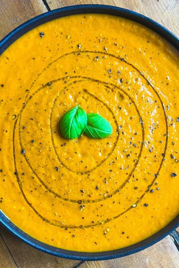 A bowl of creamy orange tomato soup garnished with a swirl of olive oil, black pepper, and two fresh basil leaves on a wooden table.