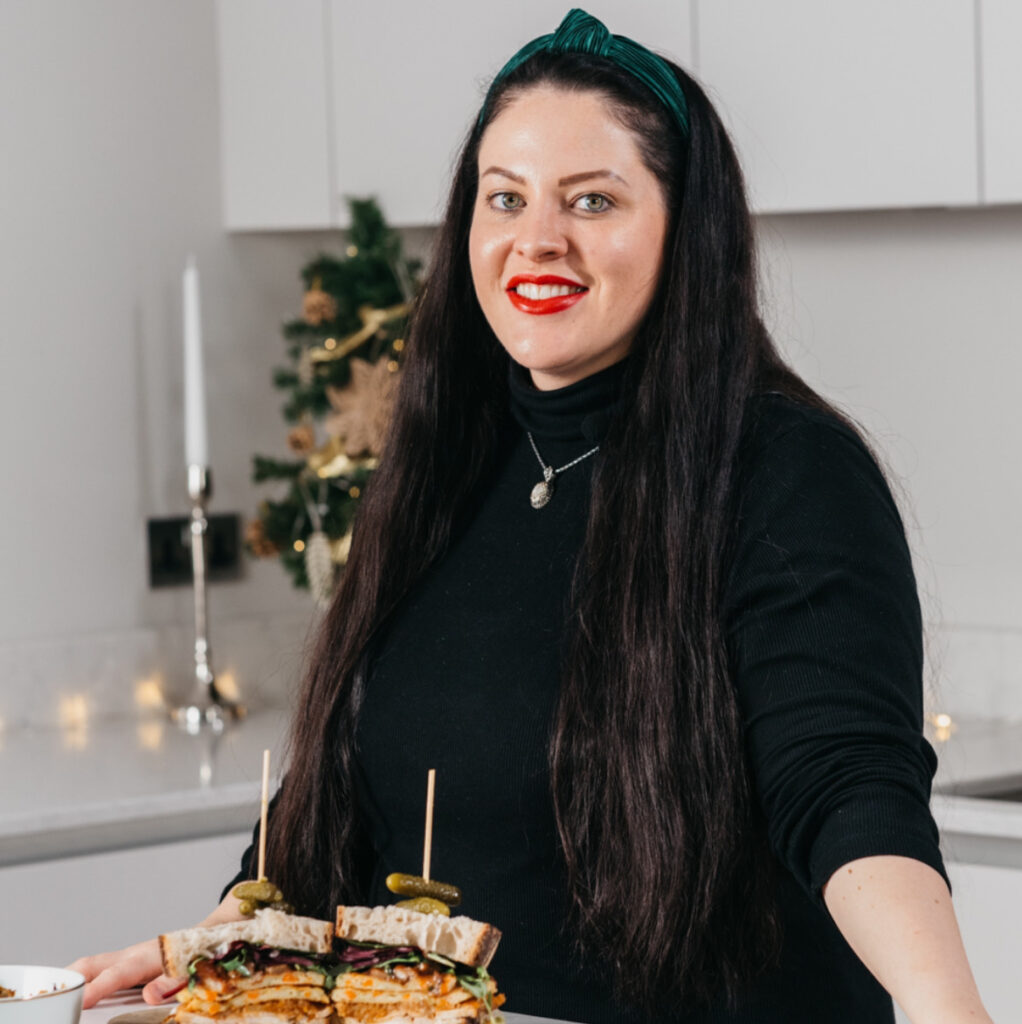 Lauren with long dark hair and red lipstick stands in a modern kitchen, smiling. In front of her is a wooden board with stacked sandwiches. A decorated Christmas tree and a lit candle are visible in the background.