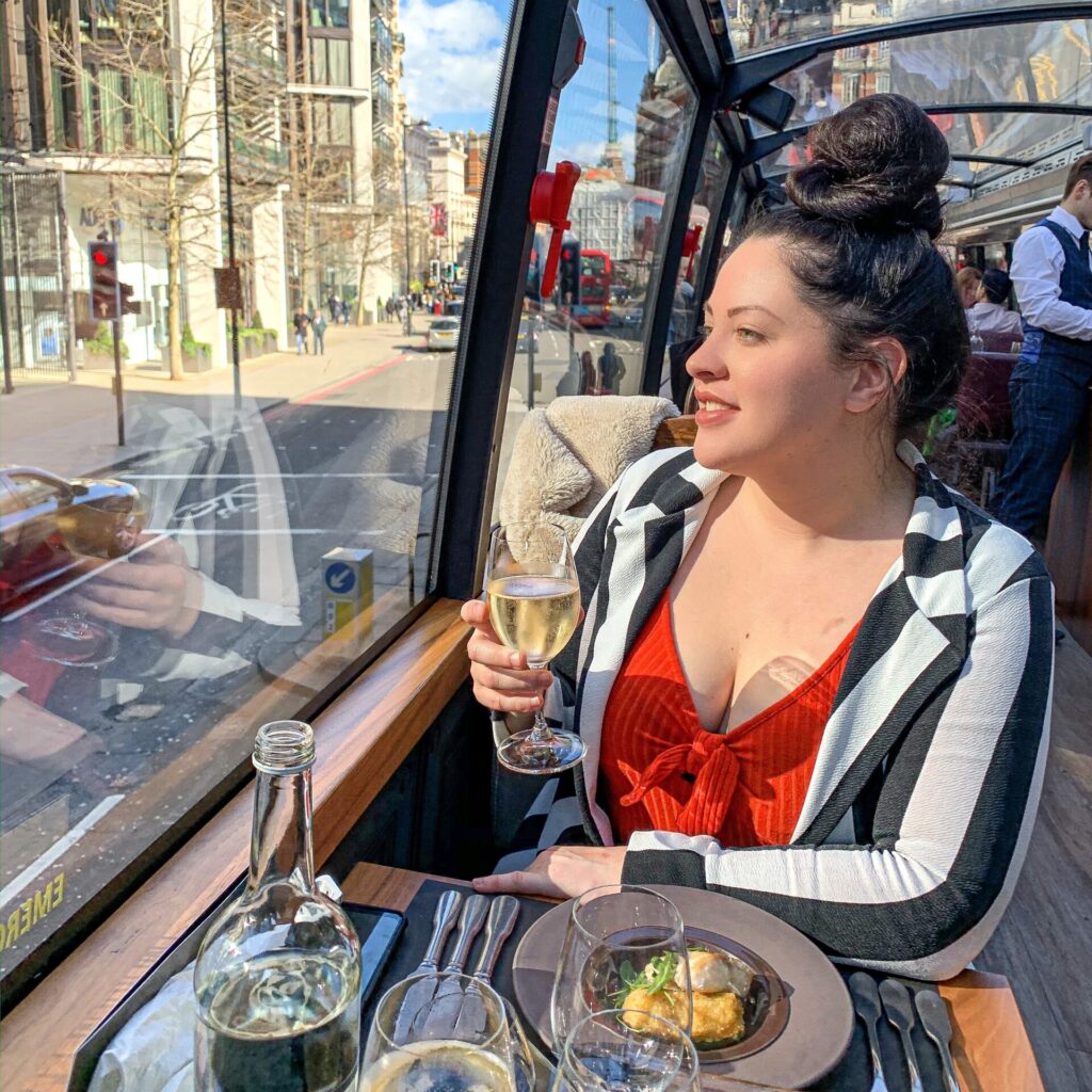 Lauren with a bun hairstyle holds a glass of white wine inside a glass-roofed restaurant. She wears a red top and black-and-white striped jacket. The scene shows a city street with tall buildings visible through the windows.