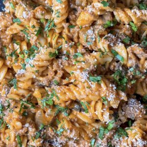 Close-up of a creamy pasta dish featuring rotini noodles mixed with a rich, savory sauce, ground meat, and garnished with chopped parsley and grated cheese. The dish appears flavorful and well-seasoned.