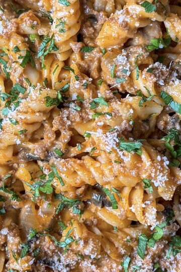 Close-up of a creamy pasta dish featuring rotini noodles mixed with a rich, savory sauce, ground meat, and garnished with chopped parsley and grated cheese. The dish appears flavorful and well-seasoned.