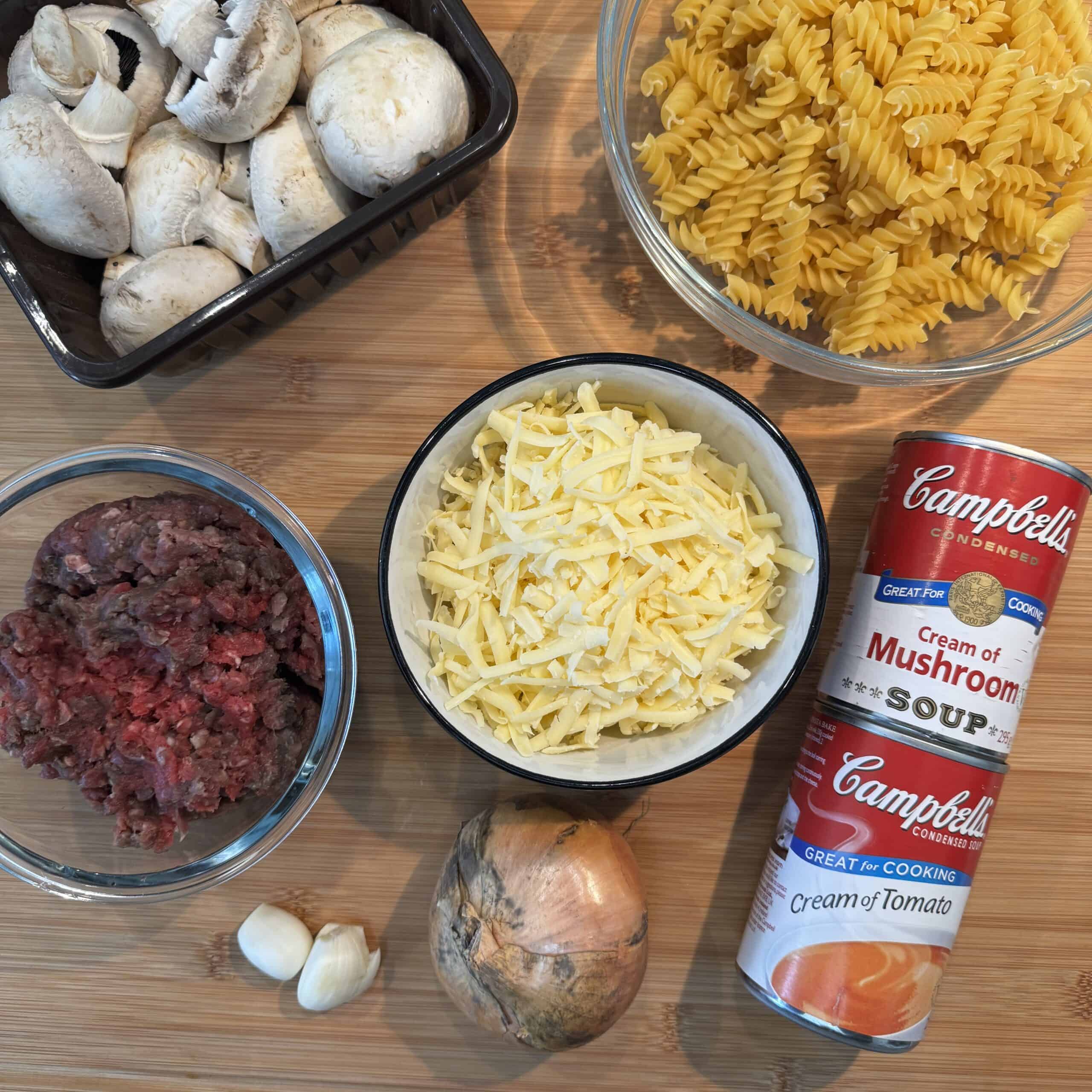Ingredients for a meal are arranged on a wooden surface: white mushrooms in a black container, uncooked spiral pasta, shredded cheese, canned cream of mushroom and tomato soup, minced meat, garlic cloves, and an onion.