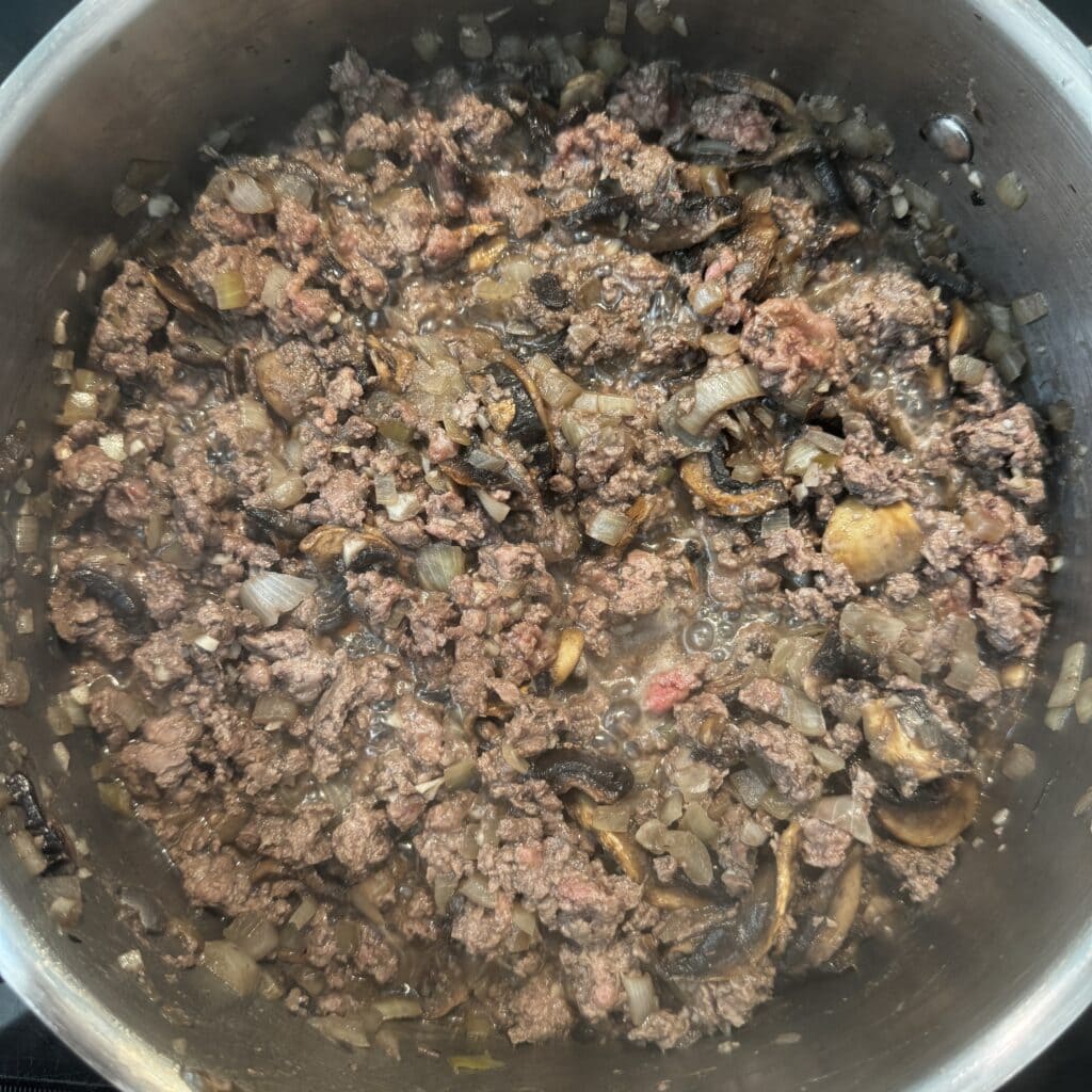 A pot containing a mixture of ground beef, chopped onions, and sliced mushrooms being cooked. The ingredients are well-mixed and appear to be sautéing, with the meat browning and the onions and mushrooms softening.