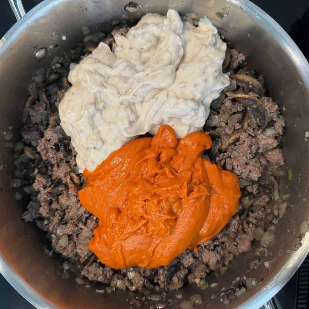 A pot containing cooked ground beef and onions, topped with dollops of cream of mushroom soup and tomato sauce, ready to be mixed or cooked further.