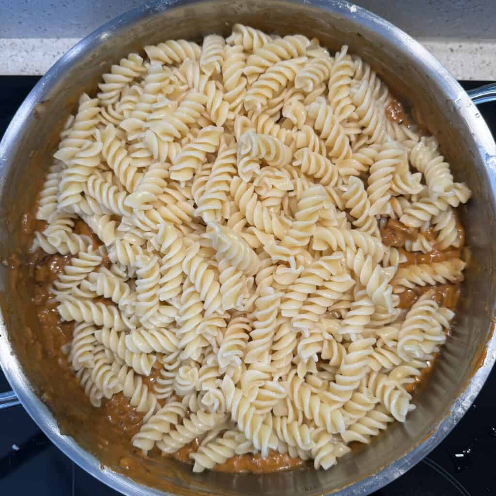 A large pan filled with cooked fusilli pasta on top of a sauce. The spiraled pasta is evenly spread across the sauce, which appears to contain a thin, orange-brown mix, possibly tomato or cheese-based. The pan is on a stove.