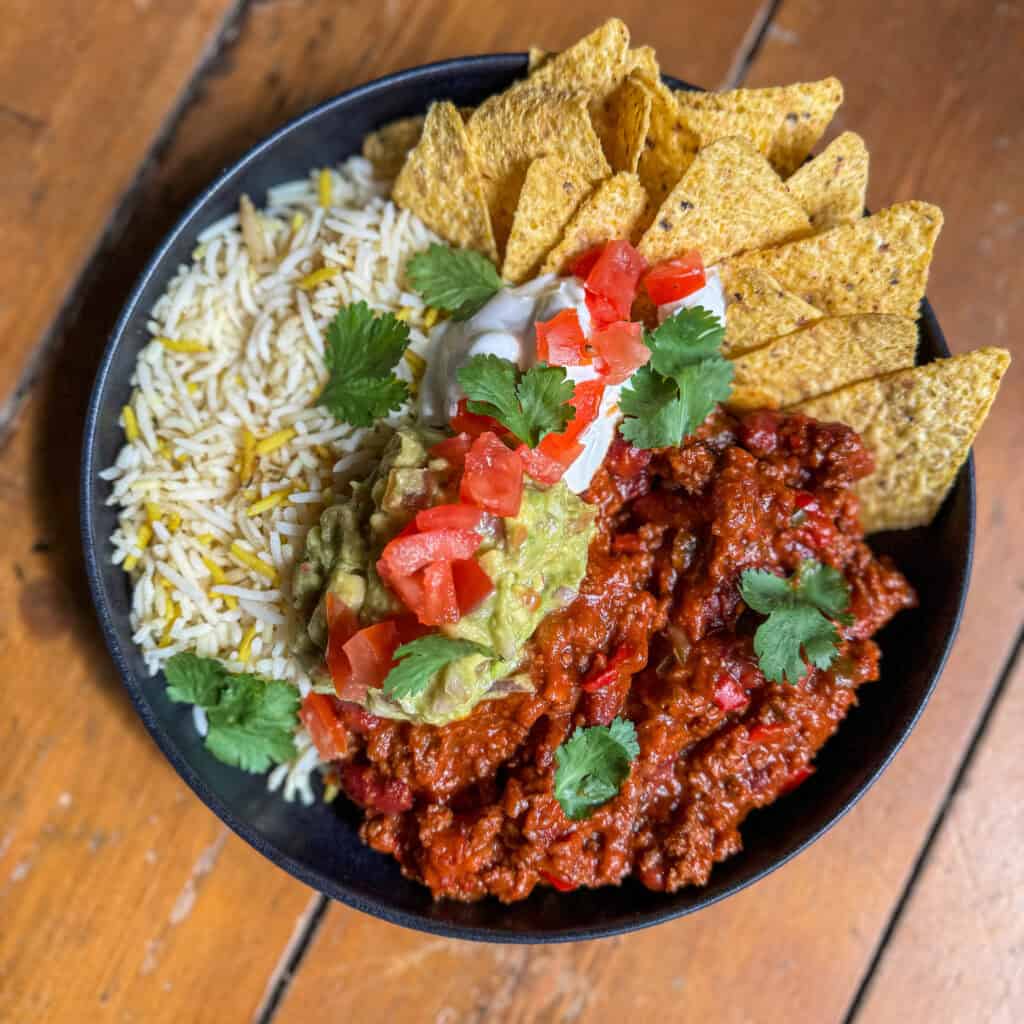 A bowl of seasoned rice, chili with minced meat, and tortilla chips garnished with tomatoes, guacamole, sour cream, and cilantro, placed on a wooden surface.