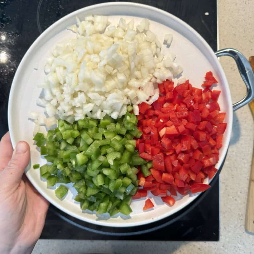 A person holds a round white plate containing diced onions, green bell peppers, and red bell peppers, arranged in separate sections. The plate is positioned above a black stove, and a countertop is partially visible.
