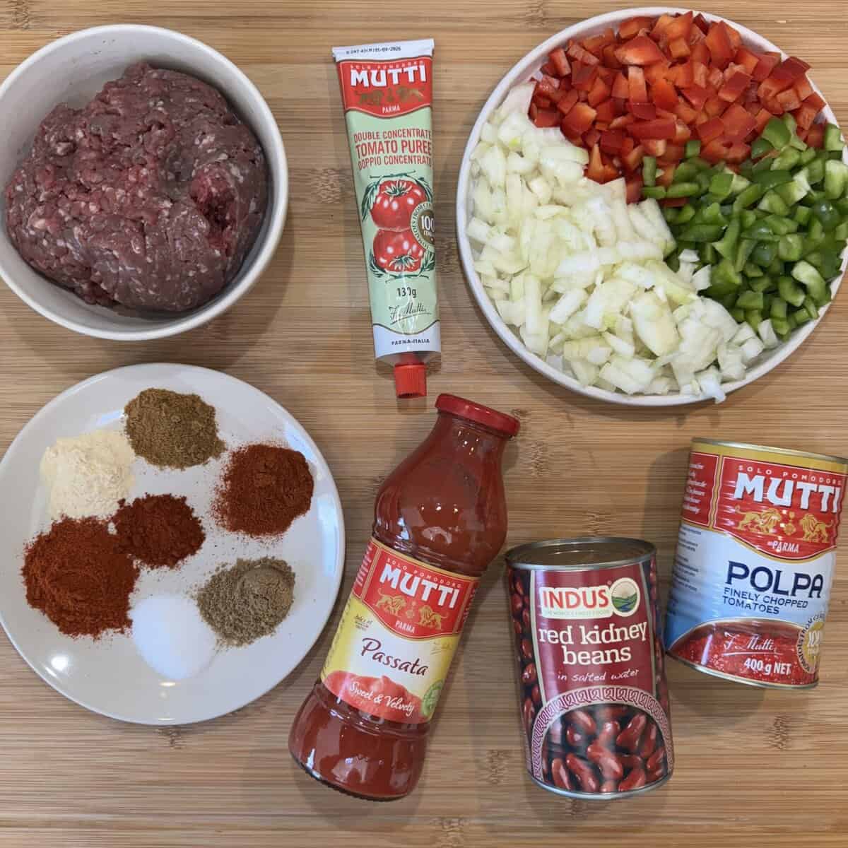 Ingredients for chili preparation on a wooden surface: a bowl of minced meat, chopped onions and peppers, tomato puree, tomato passata, canned chopped tomatoes, canned kidney beans, and a plate of various spices and salt.
