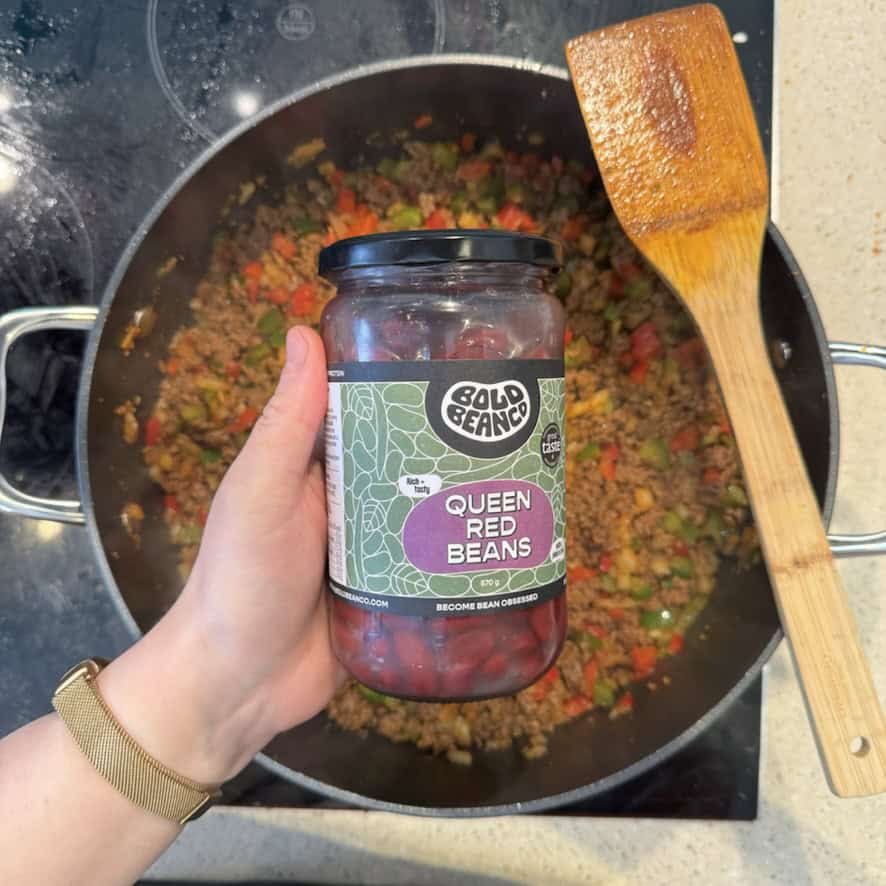 A hand holds a jar of Bold Bean Co. Queen Red Beans over a skillet on a stovetop. The skillet contains a mixture of ground meat and chopped vegetables. A wooden spatula rests on the skillets edge.
