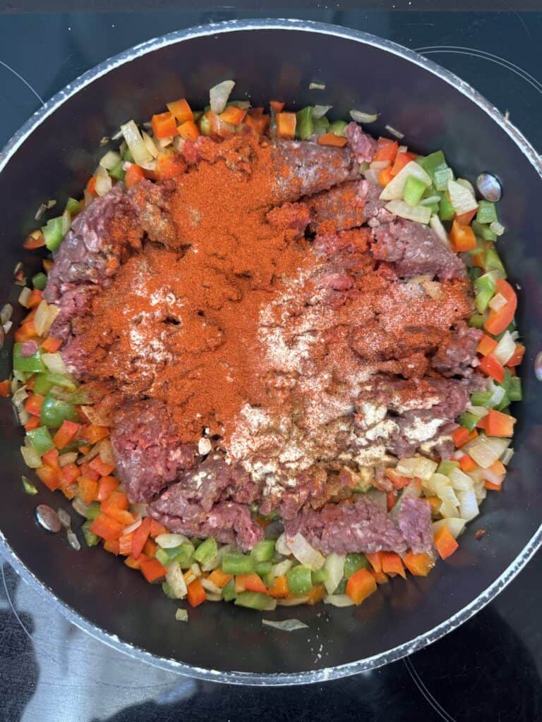 A pot of diced red and green bell peppers and onions surrounding raw minced meat, topped with red and white spices, ready to be cooked on a stovetop.