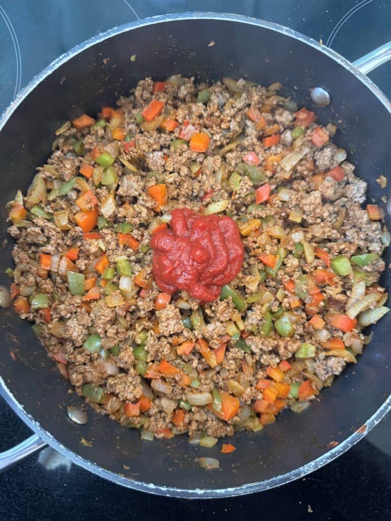 A pan filled with cooked ground meat mixed with chopped red and green bell peppers, onions, and carrots. A dollop of tomato paste is in the center. The mixture is being prepared on a stovetop.