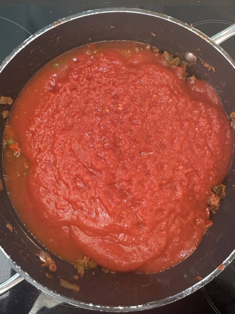 A saucepan filled with a rich tomato sauce, showing a thick, smooth texture with small visible chunks. The sauce appears to be simmering, with a vibrant red color, hinting at underlying ingredients like ground meat and vegetables.